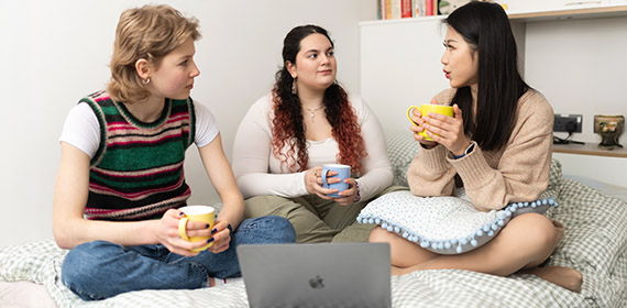 Three students are sat on a bed. In the foreground is a macbook. The students are drinking a hot